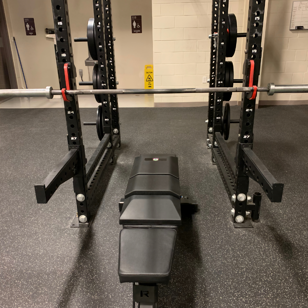 Front view of a custom-branded version of The Launch Pad® on a bench inside a black power rack, with a loaded barbell on the safety pins.